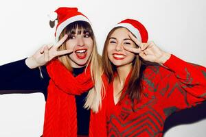 Crazy new year party mood. Two drunk laughing women having fun and posing on white background in cute masquerade hats. Red sweater and scarf. Showing signs. White teeth, bright make up. photo
