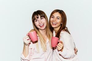 cerca arriba retrato de dos alegre blanco mujer en rosado pijama con taza de té posando en blanco antecedentes. destello retrato. foto