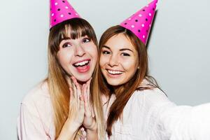 Self portrait of two smiling women in pink  paper birthday hats  on white background.  Friends wearing pink pajamas. photo