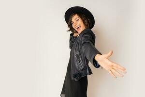 Party mood. Laughing woman  in black hat, evening dress and leather jacket  having fun and dancing on white background in studio. photo