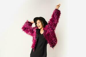 Fashionable happy model in winter pink  fur coat dancing and having fun on white background.  New year party mood. Black hat. photo