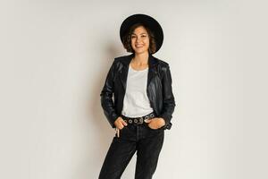 Studio fashion photo of stylish brunette woman in black hat and leather jacket posing on white bacground. White t-shirt.