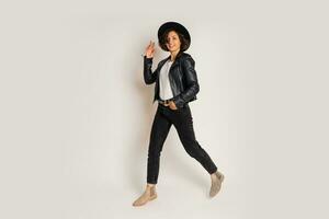 Studio fashion photo of stylish brunette woman in black hat and leather jacket posing on white bacground.  Full lenght. White t-shirt.