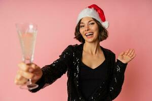 sonriente bonito mujer en Papa Noel sombrero baile, participación champú y celebrando nuevo año fiesta. posando en rosado antecedentes. elegante atuendo. foto