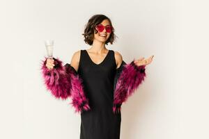 Happy  selebrating woman in stylish fur coat holding glass of shampagne , wearing pink fur coat and posing over white background. photo