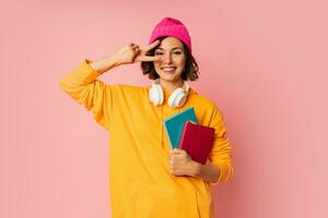 Studio photo of happy cute student with notebooks and earphones standing over pink background. Education  concept.