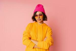 Woman in pink hat and  orange hoodie posing on pink bacground.  Stylish sunglasses. photo