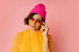 Woman in pink hat and  orange hoodie posing on pink bacground.  Stylish sunglasses. photo