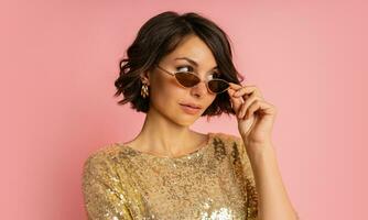 cerca arriba estudio retrato de elegante mujer en elegante lentejuela dorado vestir posando en rosado antecedentes. foto