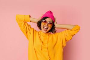 Woman in pink hat and  orange hoodie  with emotional face posing on pink bacground.  Stylish sunglasses. photo