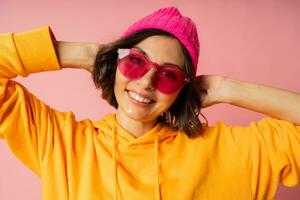 Woman in pink hat and  orange hoodie  with emotional face posing on pink bacground.  Stylish sunglasses. photo