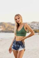 Outdoor summer image of sexy sportive blond woman posing on the beach. Wearing jeans shorts and green top. Fashion details. photo