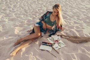 Summer lifestyle image of blond woman drawing watercolor flower by brush.  Artist sitting on the beach. Bohemian outfit. photo