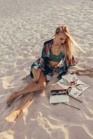 Summer lifestyle image of blond woman drawing watercolor flower by brush.  Artist sitting on the beach. Bohemian outfit. photo