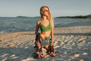 Outdoor summer image of sexy sportive blond woman posing on the beach. Wearing jeans shorts and green top. Fashion details. photo