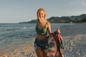 Outdoor summer image of sexy sportive blond woman posing on the beach. Wearing jeans shorts and green top. Fashion details. photo