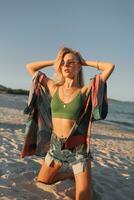 Outdoor summer image of sexy sportive blond woman posing on the beach. Wearing jeans shorts and green top. Fashion details. photo