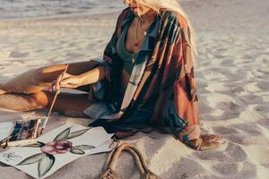Summer lifestyle image of blond woman drawing watercolor flower by brush.  Artist sitting on the beach. Bohemian outfit. photo