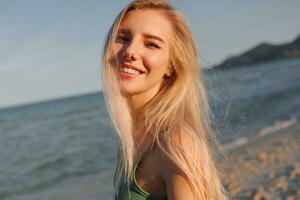 Outdoor summer image of sexy sportive blond woman posing on the beach. Wearing jeans shorts and green top. Fashion details. photo