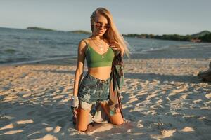 Outdoor summer image of sexy sportive blond woman posing on the beach. Wearing jeans shorts and green top. Fashion details. photo