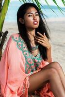 tropical verano retrato de bonito asiático joven mujer en brillante ropa de playa, boho vestir con bordado y borla relajante en increíble playa, sentado en blanco arena cerca palma árbol, mar antecedentes. foto