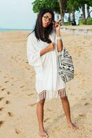 Seductive Asian woman in black top and jeans shorts , beach cover up with boho bag posing on tropical beach. Bohemian girl enjoying weekends. Perfect body.Jewelry ,bracelet and necklace. photo