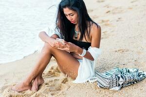 Young pretty Asia woman holding and using mobile phone testing on tropical beach. Wearing stylish white boho cover up and trendy accessories . Red lips. photo