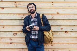 Close up image of hipster stylish man trying to use retro film camera ,posing on wooden wall. Wearing leather jacket, backpack and plaid shirt. photo