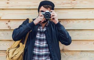Handsome hipster man with beard, in stylish hat and glasses posing with retro camera in hands  and backpack stands on wooden  background. photo
