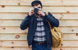 Close up image of hipster stylish man trying to use retro film camera ,posing on wooden wall. Wearing leather jacket, backpack and plaid shirt. photo