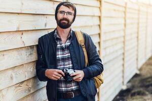 Handsome hipster man with beard, in stylish hat and glasses posing with retro camera in hands  and backpack stands on wooden  background. photo