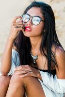 Close up lifestyle portrait of stylish Asian woman with red lips. She is sitting on sunny tropical beach. Boho accessories.Jewelry ,bracelet and necklace. photo