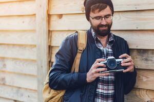 Close up image of hipster stylish man trying to use retro film camera ,posing on wooden wall. Wearing leather jacket, backpack and plaid shirt. photo