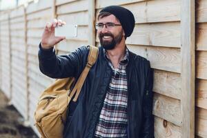 hipster hombre haciendo selfie en teléfono inteligente por móvil teléfono en madera pared antecedentes. vistiendo elegante negro sombrero, cuero chaqueta y mochila. foto