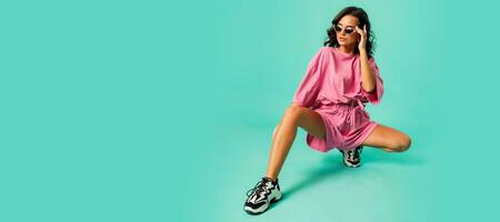 Indoor photo of  fashionable  woman posing on turquoise  background. Pretty  tan female in pink  clothes and unusual sneakers.