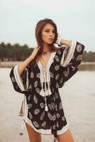 hermosa asiático mujer en tropical bohemio vestir posando en el playa. vistiendo pendientes con pluma, collar y esposas. foto