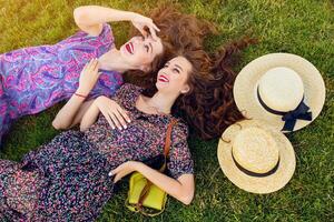 Outdoor lifestyle  image of two best friends in colorful boho dress and curly hair  laying on the green grass and talking about. photo