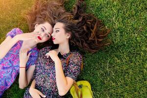 Outdoor lifestyle  image of two best friends in colorful boho dress and curly hair  laying on the green grass and talking about. photo