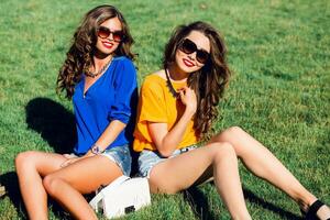 Two pretty girls  in bright summer clothes posing  on grass  and enjoying  sunny day together . Bright colors. photo