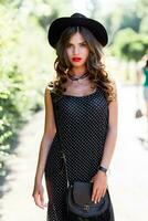 verano Moda retrato de r elegante mujer con Perfecto ondulado pelo en elegante elegante negro sombrero y brillante hacer arriba posando en el parque. calle estilo. foto