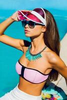 estilo de vida retrato de bronceado hermosa mujer en rosado bikini y Gafas de sol sentado cerca nadando piscina con Fresco cóctel. increíble sensual niña con Perfecto Delgado deporte cuerpo posando exterior. foto