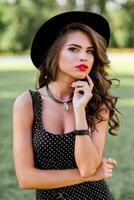 verano Moda retrato de r elegante mujer con Perfecto ondulado pelo en elegante elegante negro sombrero y brillante hacer arriba posando en el parque. calle estilo. foto