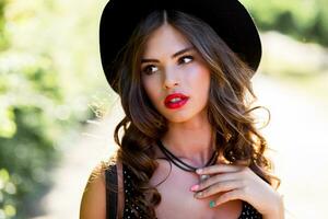 Summer fashion  portrait of r elegant woman with  perfect wavy  hair   in  stylish elegant black hat and bright make up posing in the park.  Street style. photo