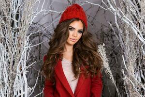 cerca arriba retrato de elegante niña en escarchado invierno parque .hermoso joven mujer en rojo de punto sombrero, ondulado increíble peinado , lleno labios y brillante hacer arriba. foto