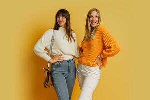 Studio photo of two pretty woman in cozy sweaters posing over yellow background.  Autumn and winter fashion trends.