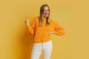 Happy  blond  woman  in orange  stylish autumn sweater posing over yellow background in studio.  Showing sign ok. photo