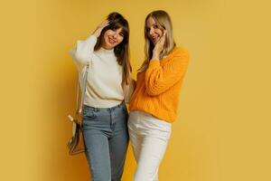 Studio photo of two pretty woman in cozy sweaters posing over yellow background.  Autumn and winter fashion trends.
