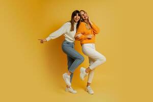 Two pretty women , best friends in stylish autumn  casual clothes having fun  over yellow background in studio. Full lenght. photo