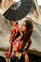 Fashion photo of elegant asian woman in silk kimono  holding fan and posing over rocks on the beach.