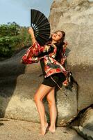 Beautiful japanese  woman in stylish  kimono  with large fan and professional make up posing over rocks. Full lenght. photo